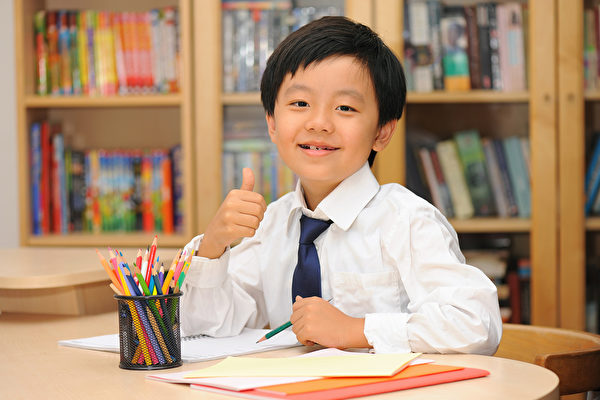 Smart Asian schoolboy sitting at a desk with thumbs up