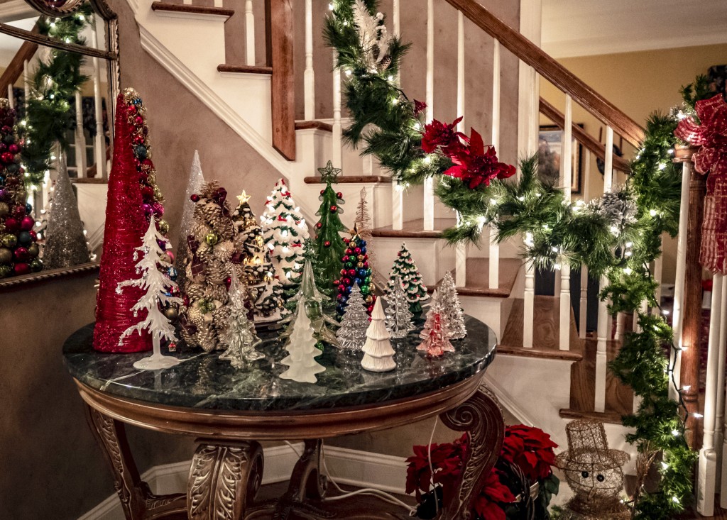 Decorative lighted Christmas trees decorate table with lighted garland on stair railing in background.