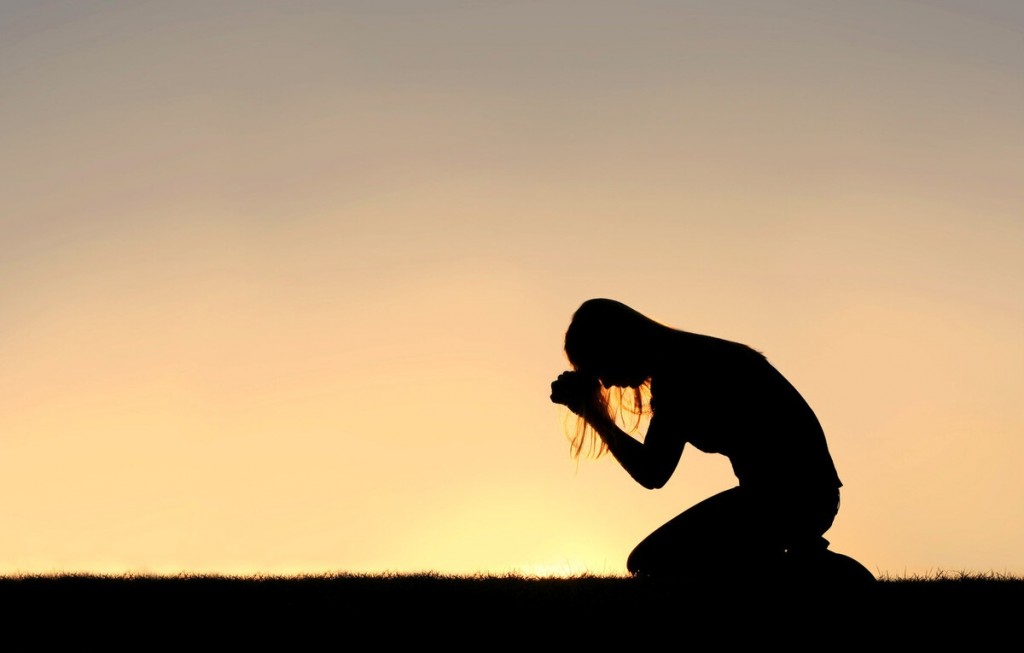 A silhouette of a young Christian woman is bowing her head in prayer, and desperation outside during sunset.