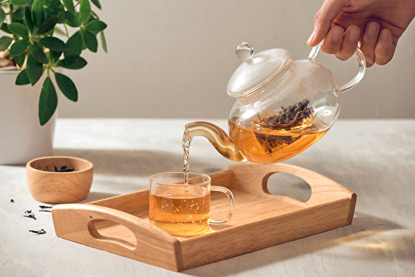 A,Hand,Pouring,Tea,From,Glass,Teapot,On,Wooden,Serving