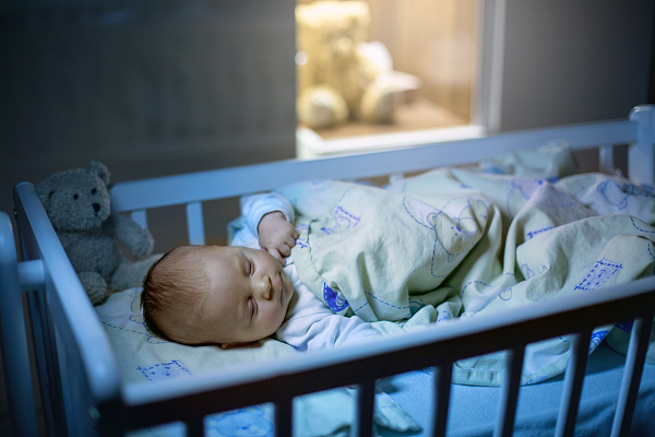 Adorable newborn baby boy, sleeping in crib at night. Little boy in white pajamas taking a nap in dark room. Bedroom interiоr with lamp and teddy bear in the background